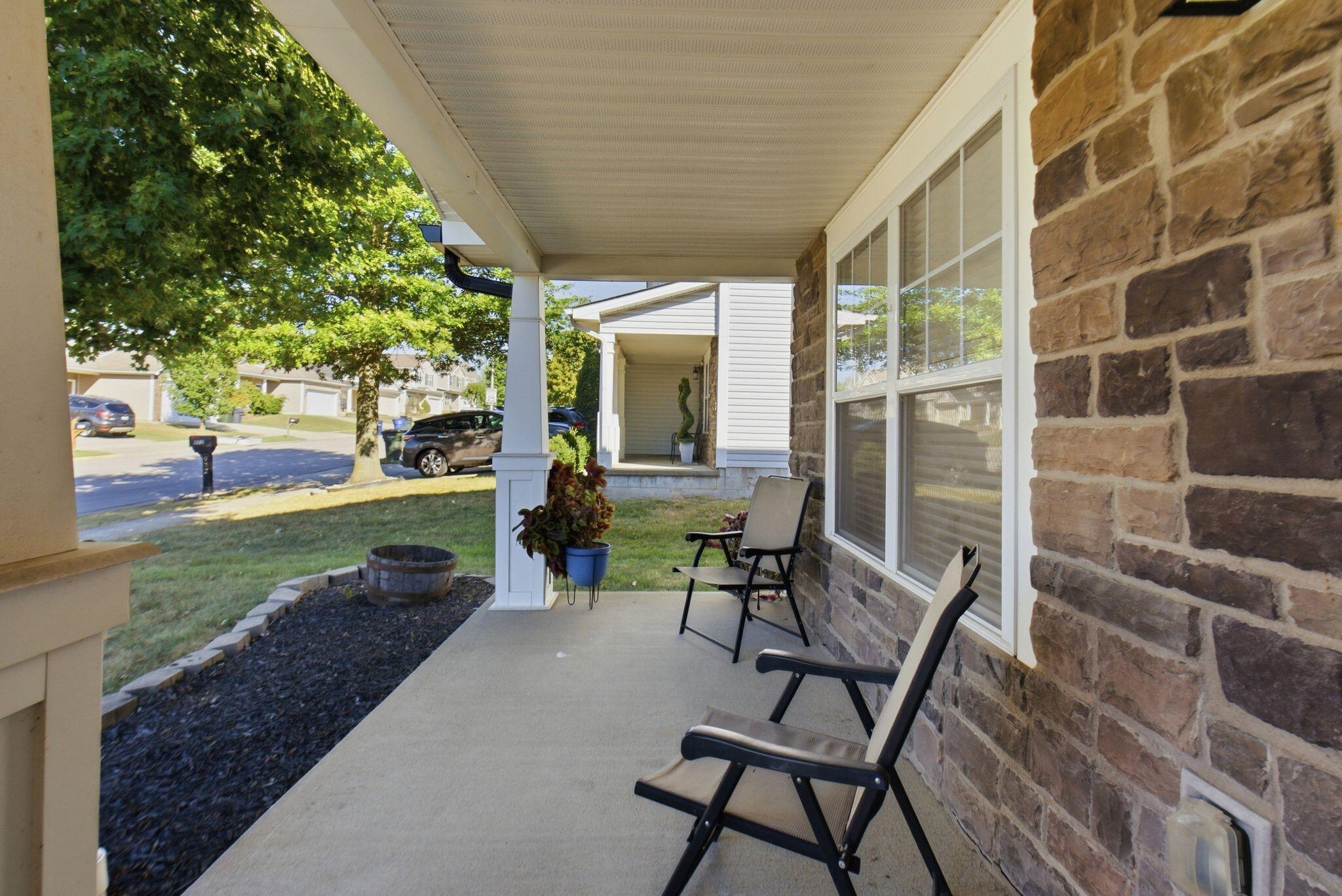 Covered front porch with stone walls, two chairs, and potted plants beside the lawn.