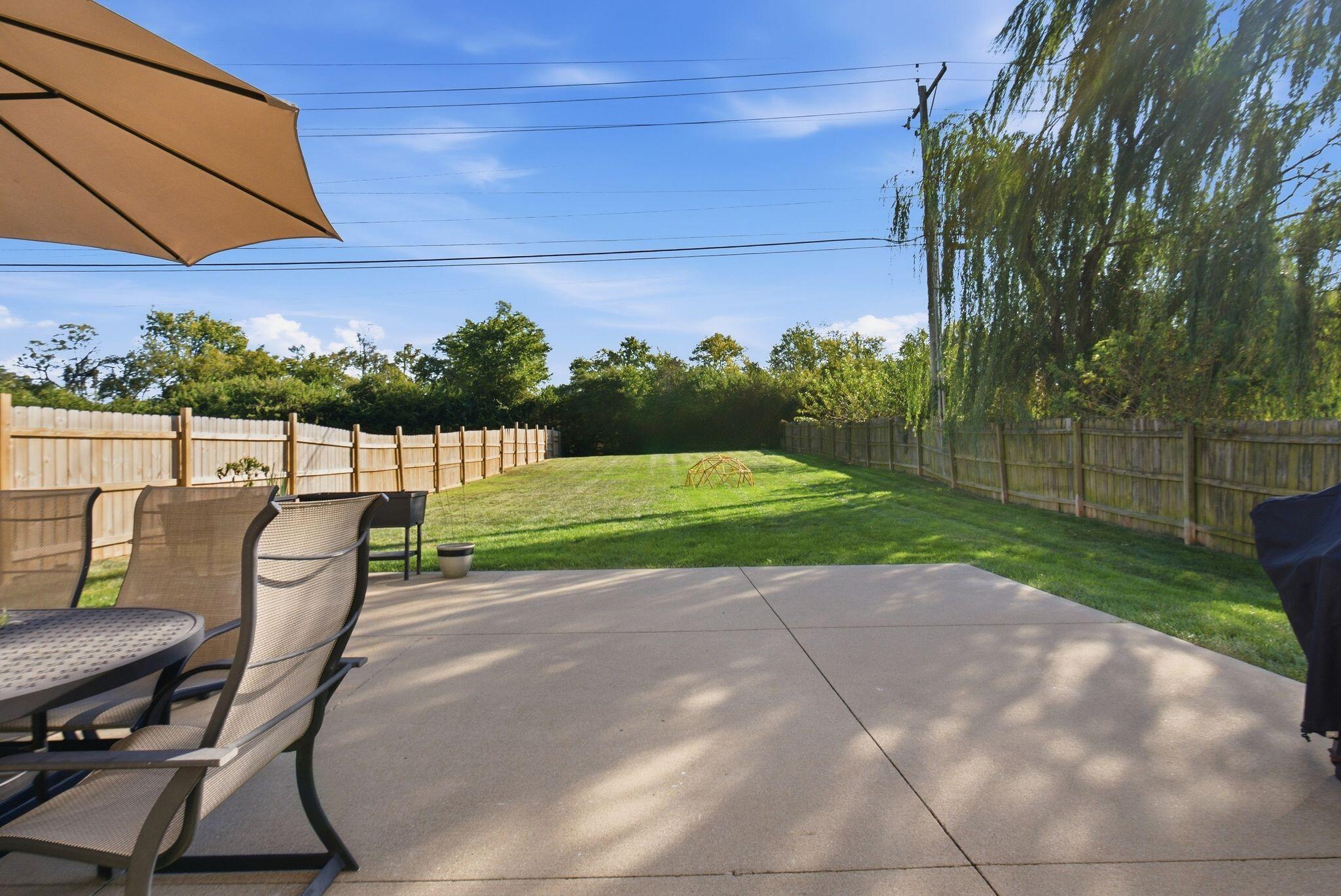 Furnished concrete patio overlooking a long, fenced grassy backyard beneath a large weeping tree.