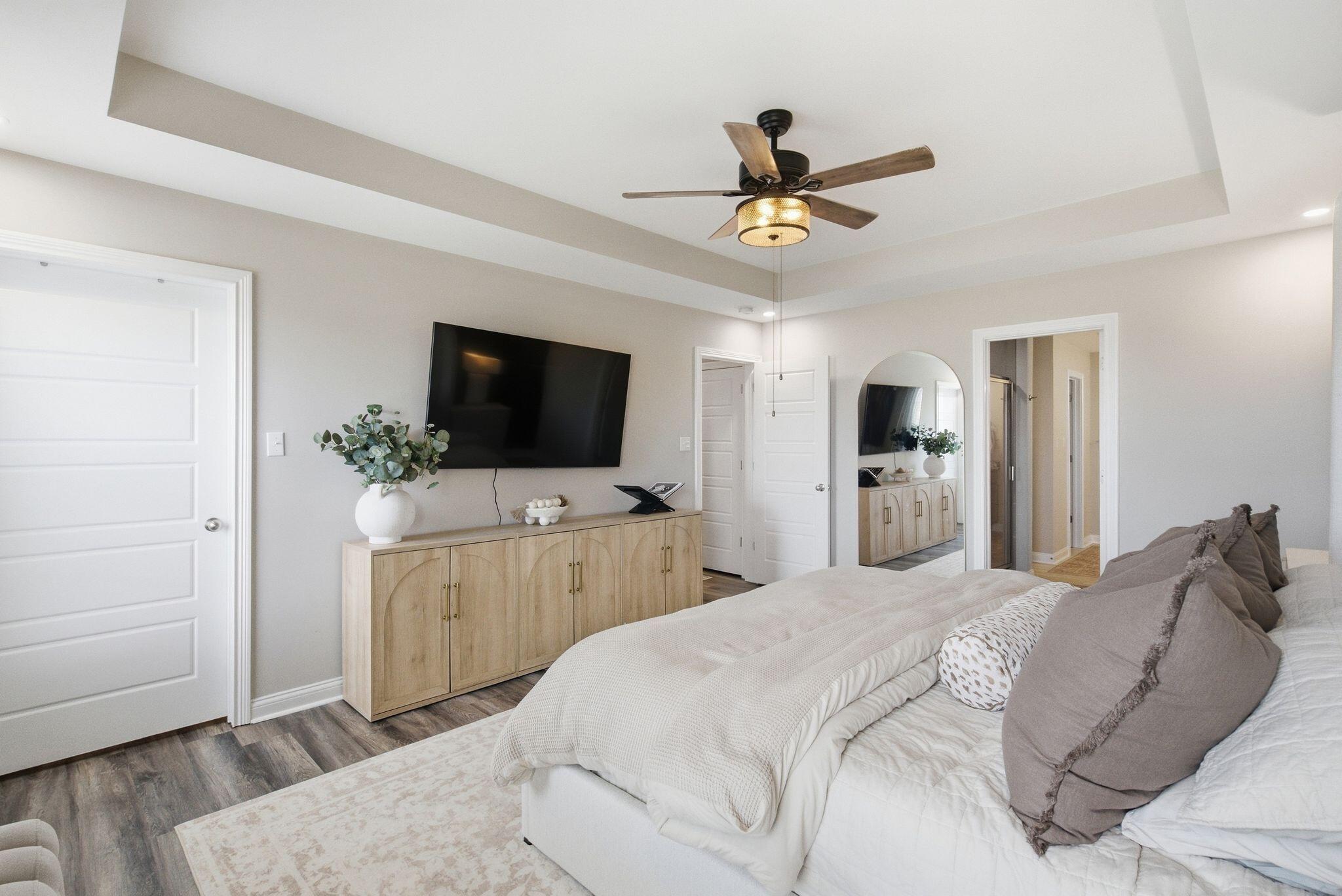 Bedroom with a tray ceiling, ceiling fan, wall-mounted TV, and neatly made bed.