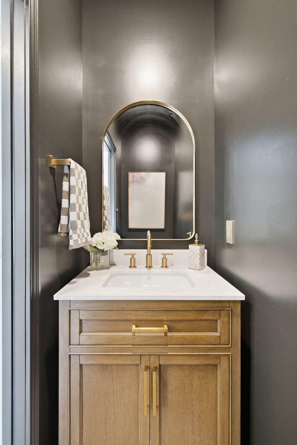 Bathroom vanity with a wood cabinet, white countertop, brass fixtures, and arched mirror.