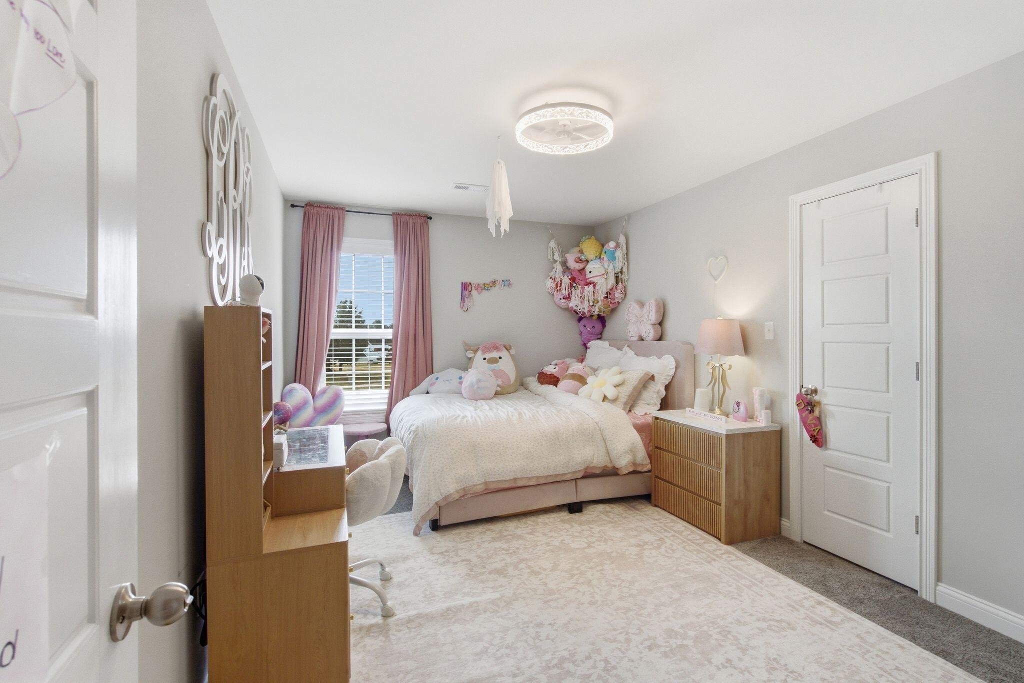 A furnished bedroom with a bed, pink curtains, plush toys, and a ceiling light.