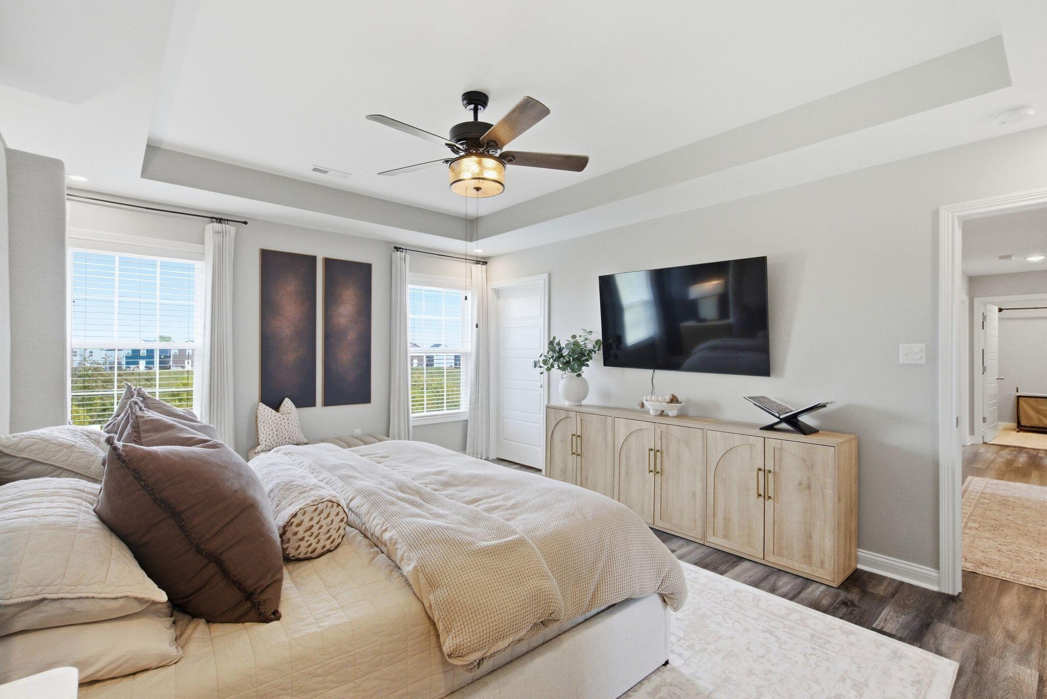 Bright furnished bedroom with a tray ceiling, ceiling fan, and wall-mounted television.