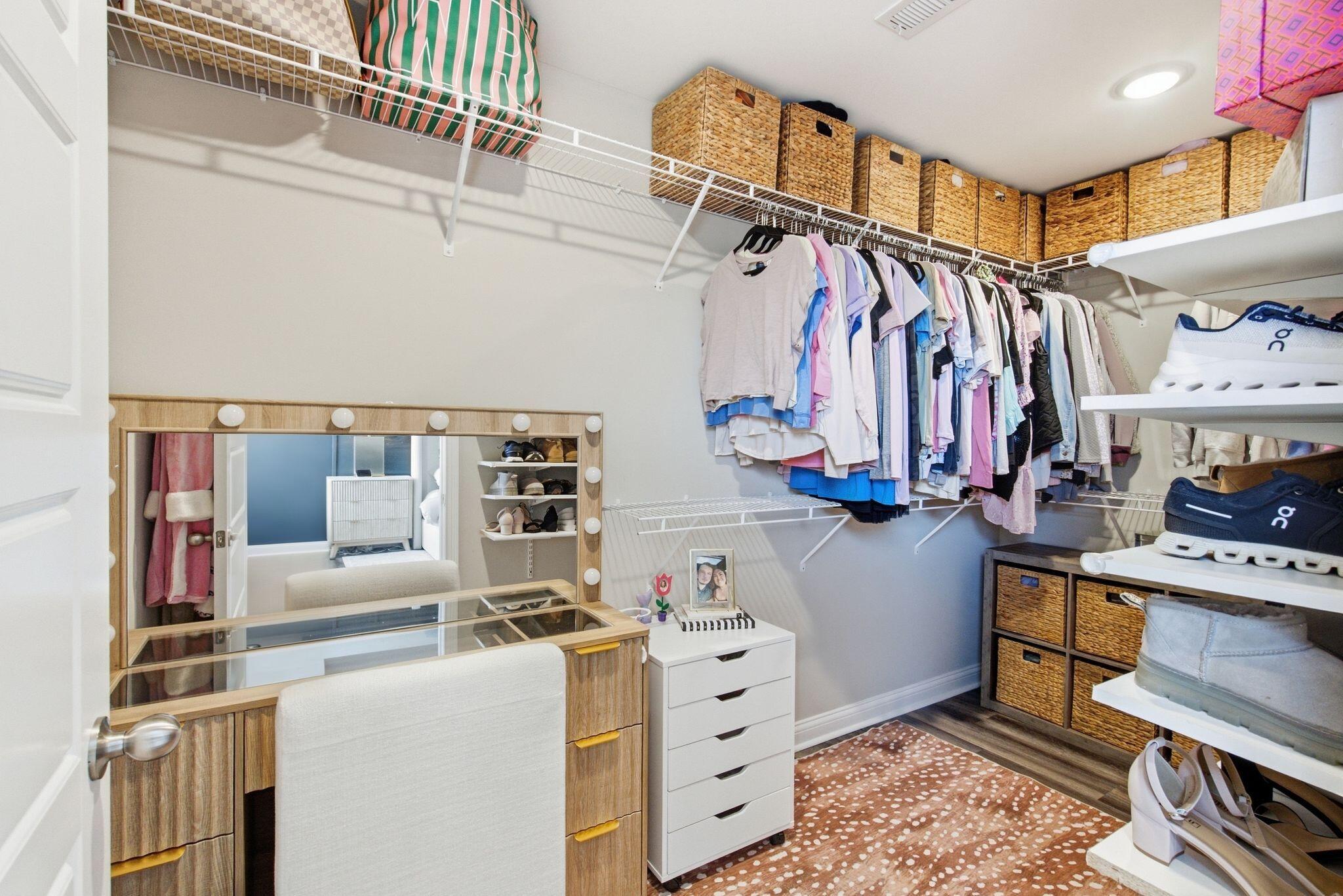 Walk-in closet with hanging clothes, shoe shelves, woven baskets, and a vanity mirror.