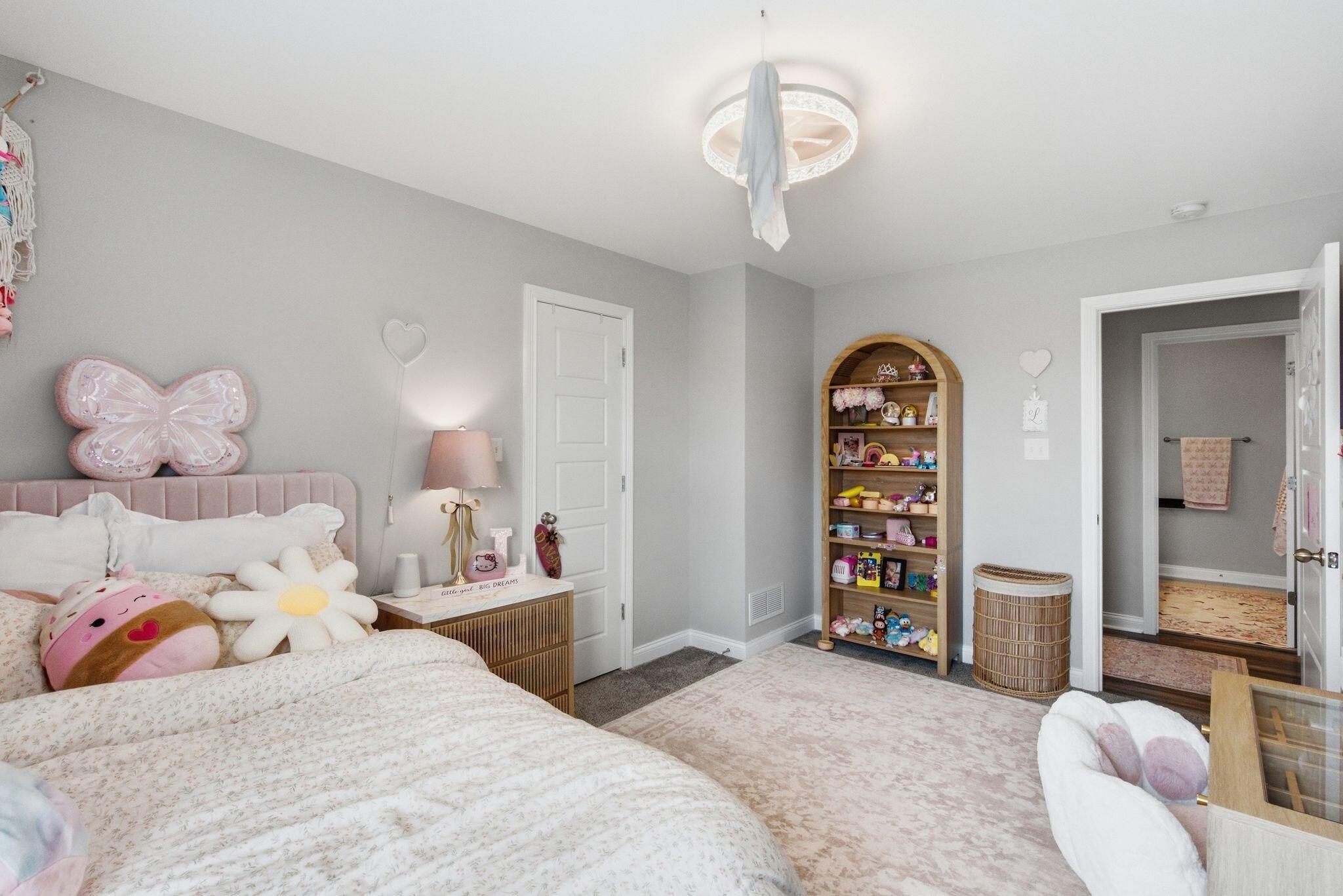 Child’s bedroom with a pink upholstered bed, butterfly cushion, toy-filled bookshelf, and adjoining bathroom.