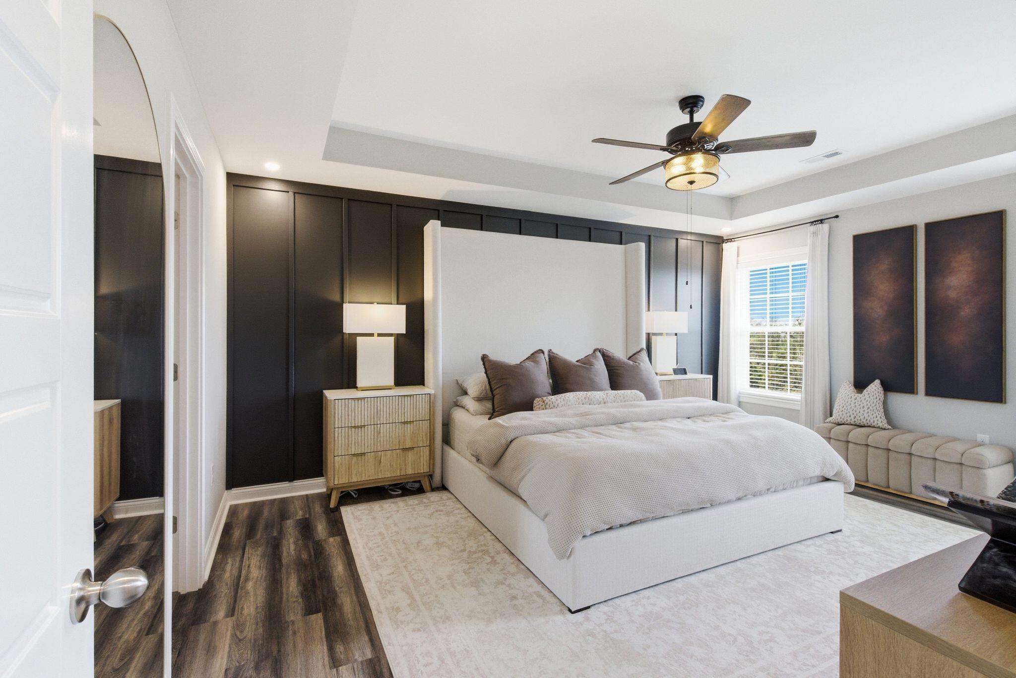 Staged bedroom with a white upholstered bed, dark paneled accent wall, and ceiling fan.