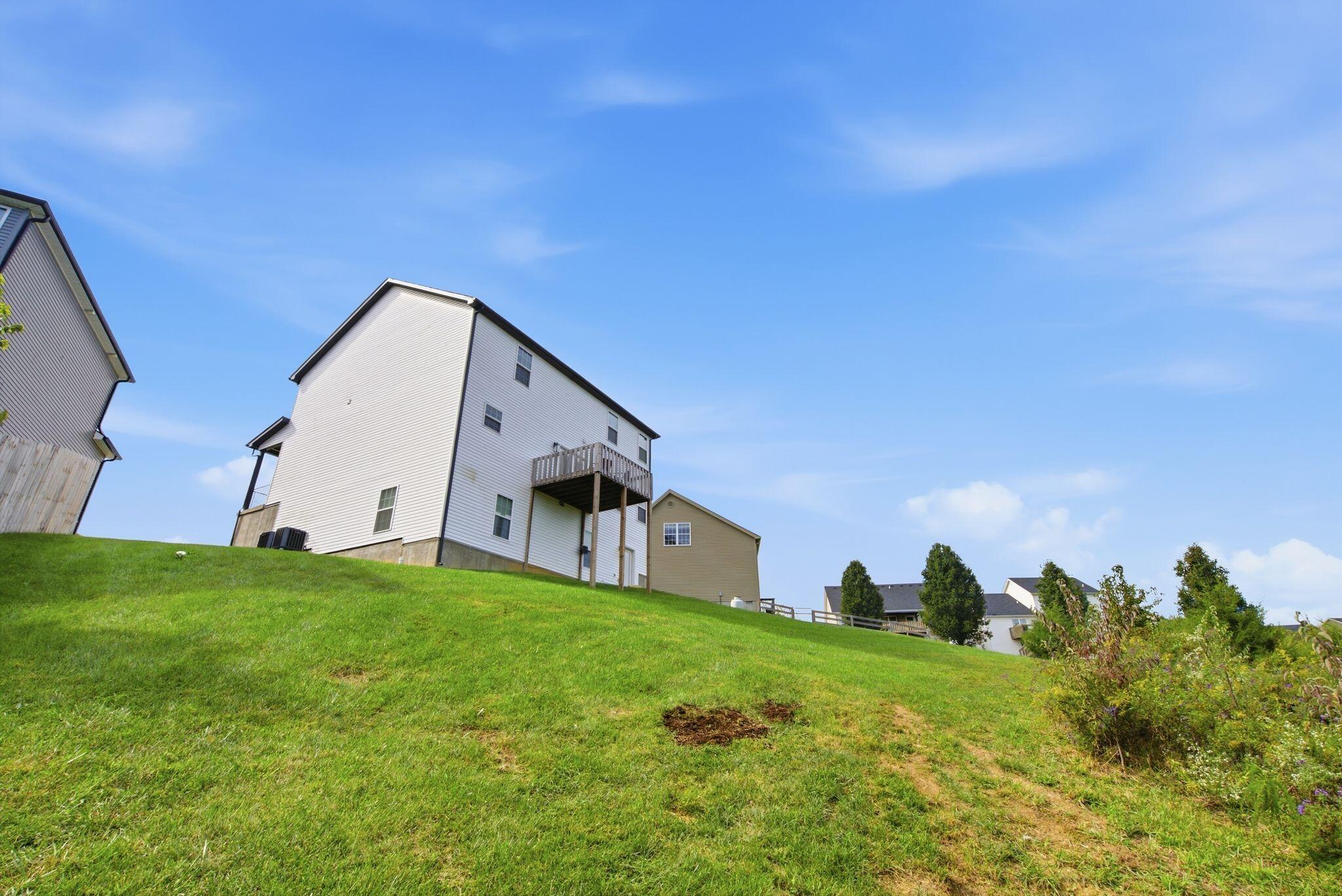 Rear view of a two-story white-sided house with a raised deck above a sloping lawn.