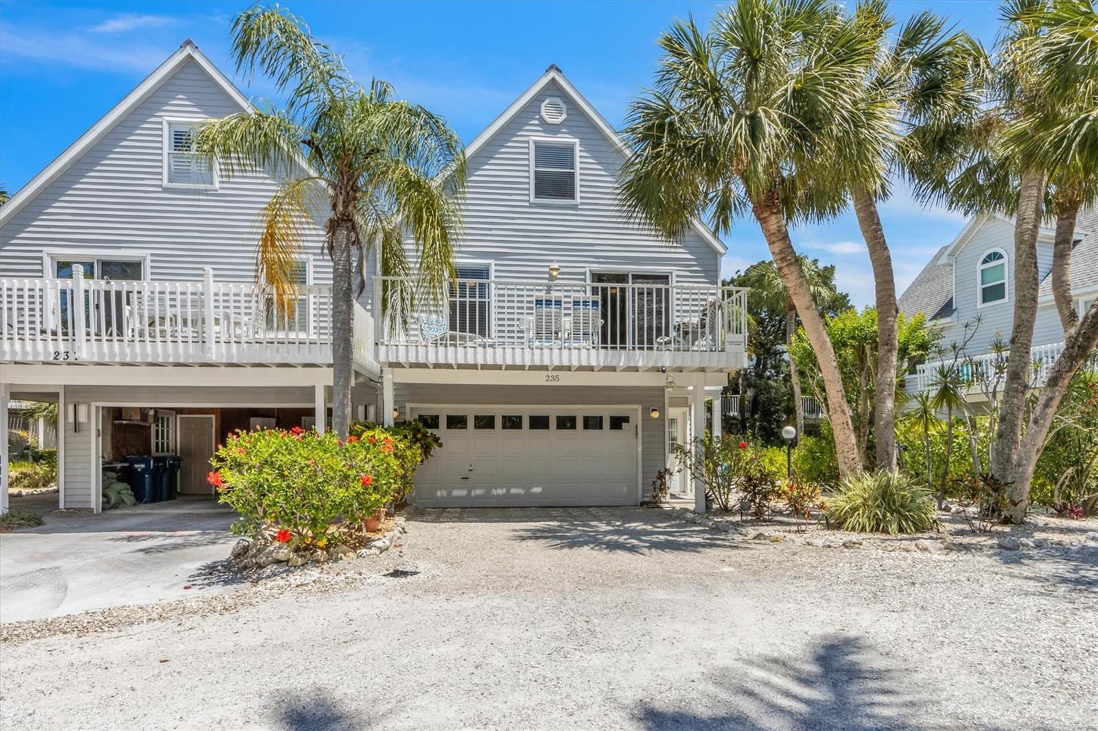 Gray coastal home with a raised white deck, double garage, palm trees, and gravel driveway.
