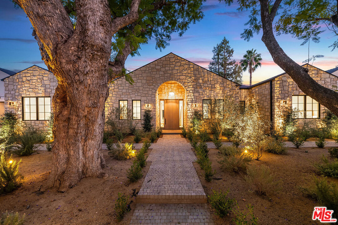 Stone house at dusk with an arched entry, illuminated windows, and a brick walkway framed by mature trees.