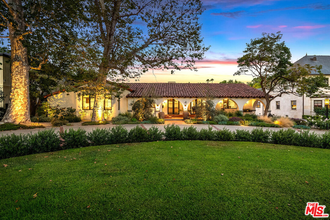 Stucco home with a red tile roof and arched entryways, warmly lit at dusk beneath mature trees.