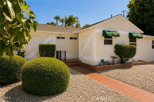 Single-story cream stucco home with green awnings, rounded shrubs, and a brick walkway across the gravel yard.