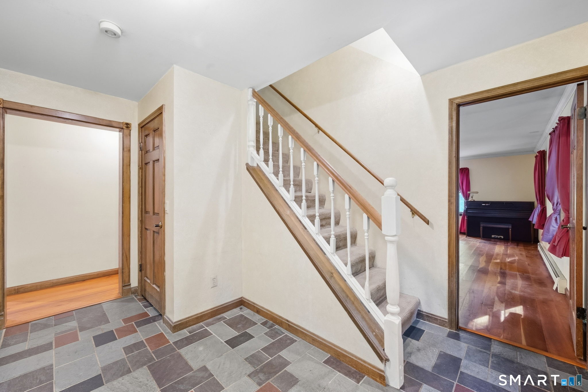 Foyer with carpeted staircase, white balusters, slate tile floor, and a piano visible in the adjoining room.
