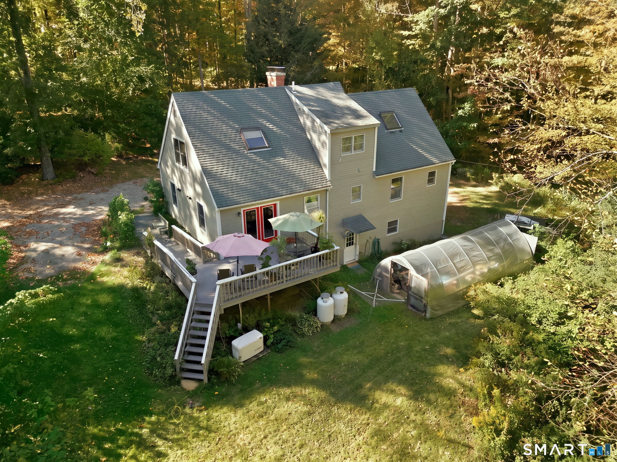 Aerial view of a beige house with a raised deck and greenhouse in a wooded yard.