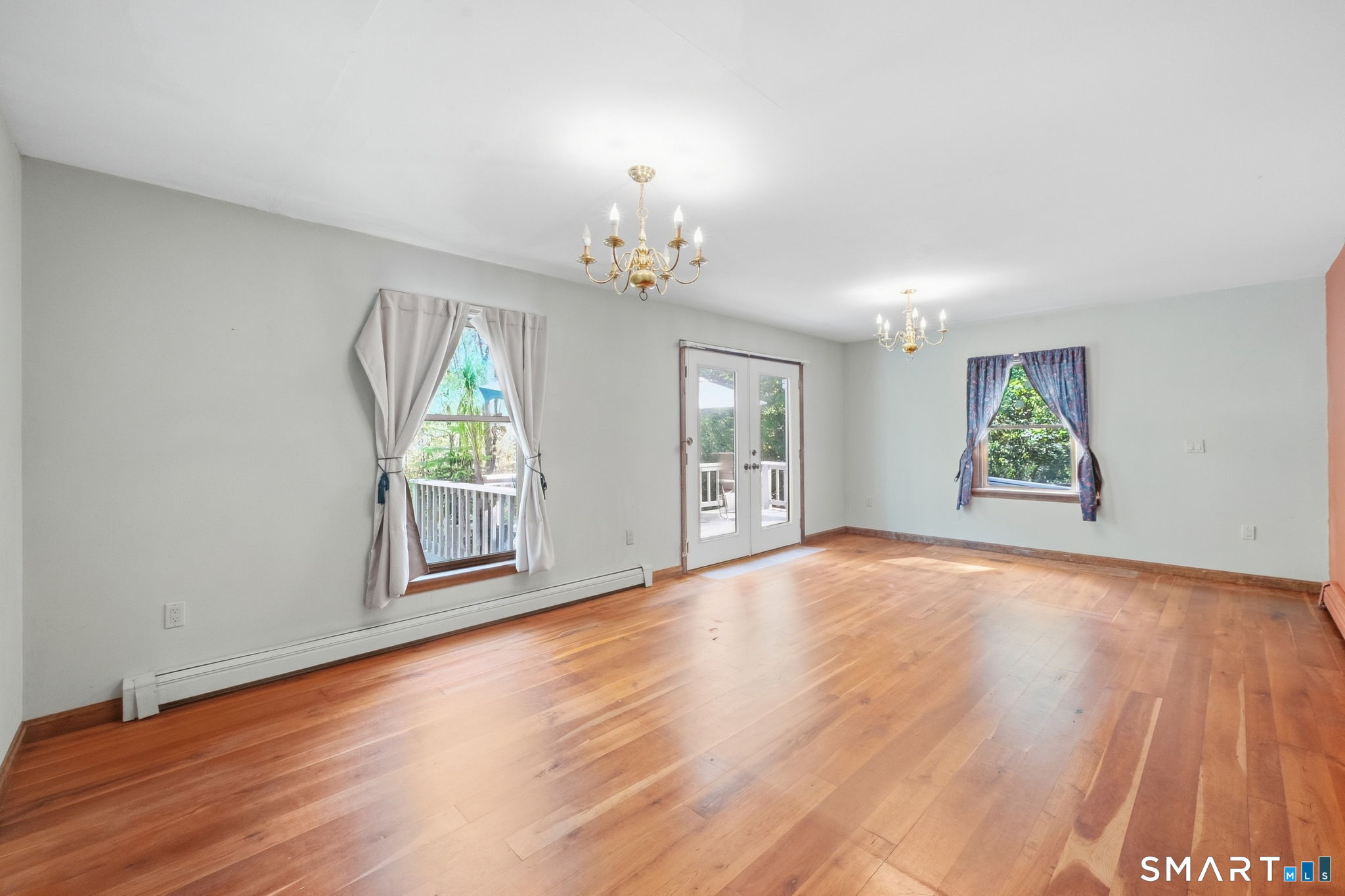 Empty room with polished hardwood floors, two chandeliers, and French doors opening to a deck.