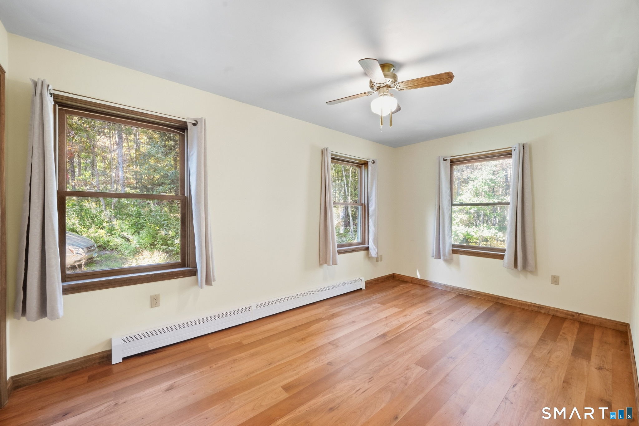 Empty room with hardwood floors, three curtained windows, a ceiling fan, and baseboard heating.