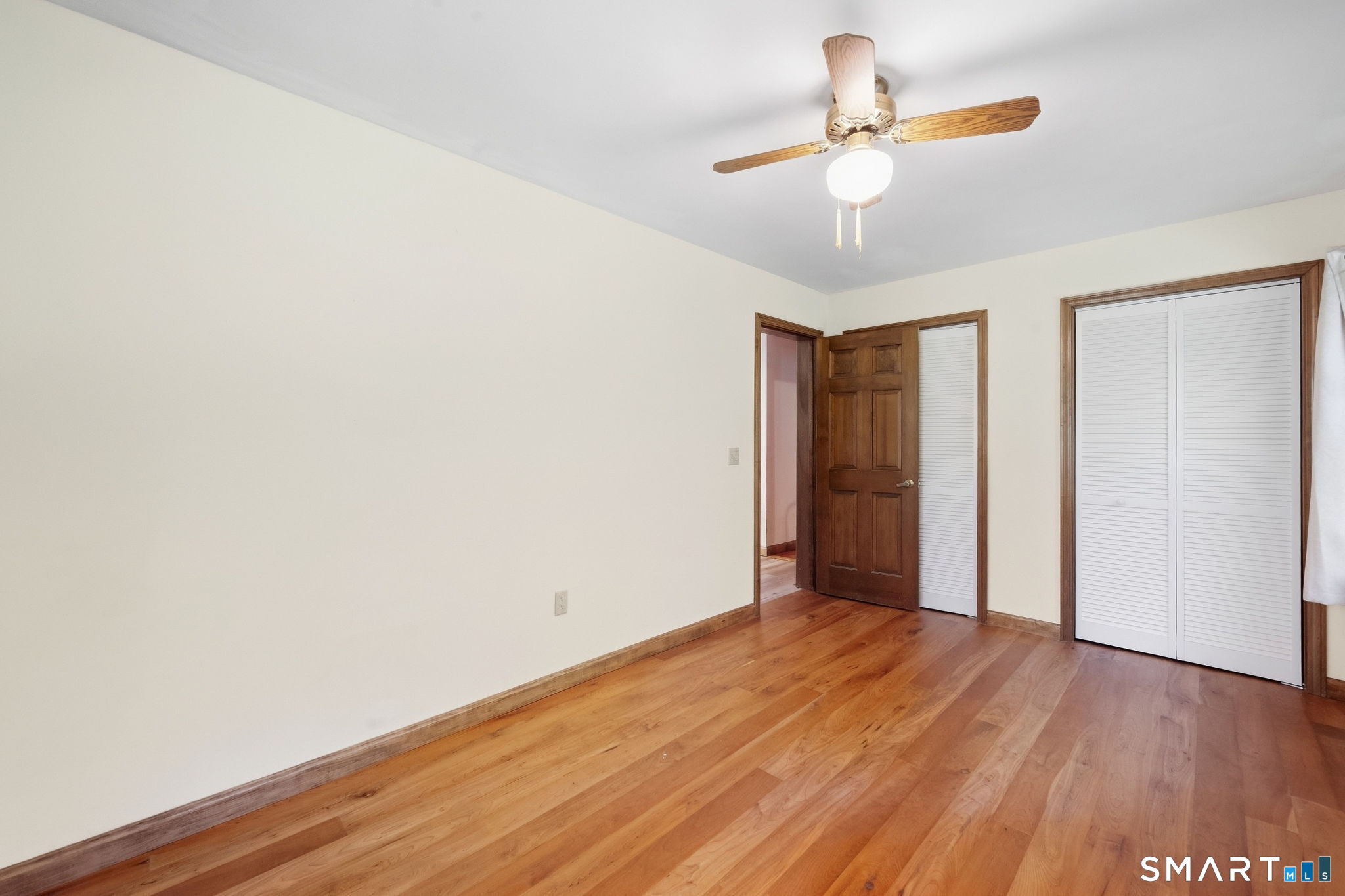 Empty bedroom with hardwood floors, a ceiling fan, and louvered closet doors.