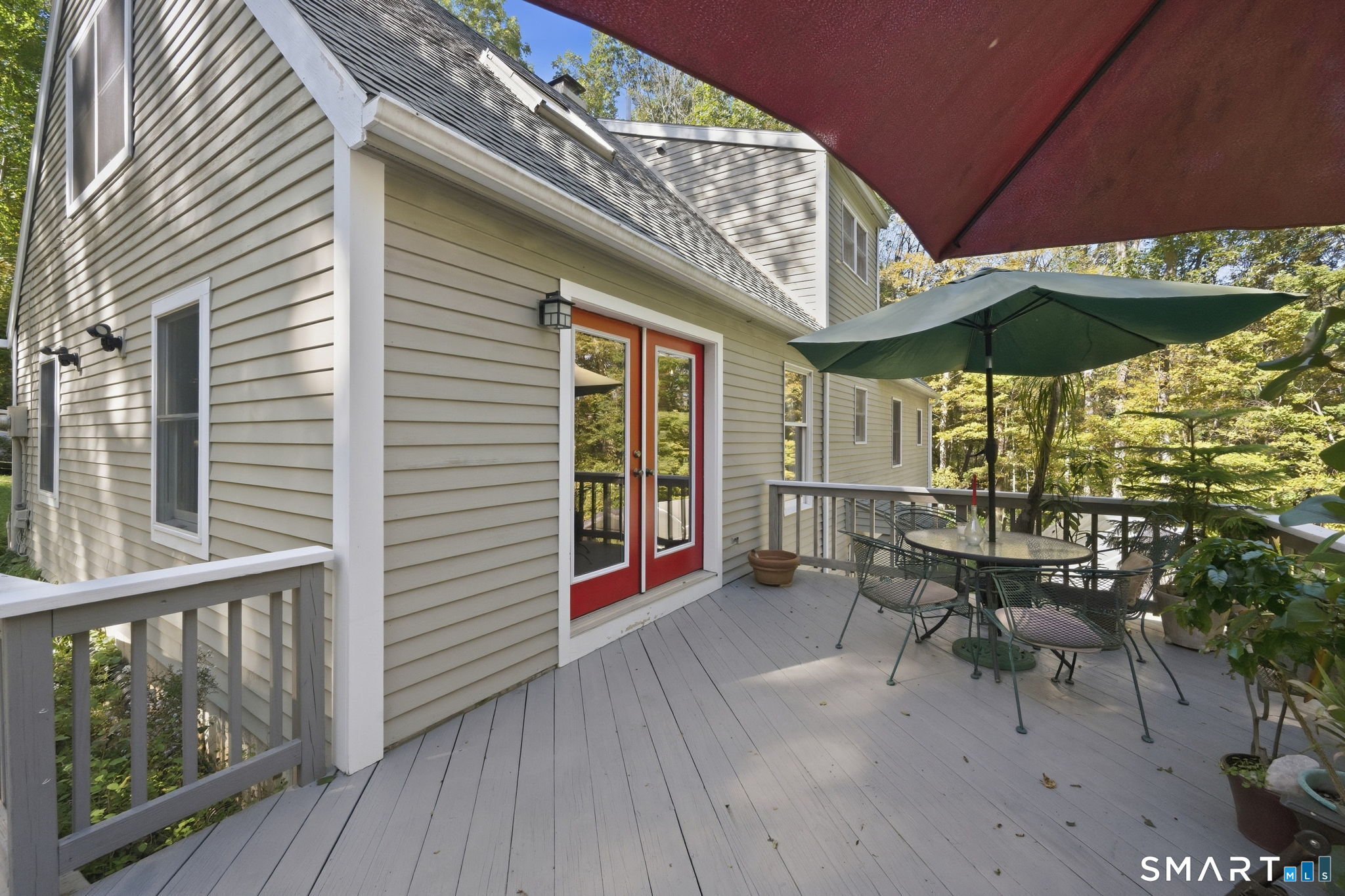Raised deck with red French doors, patio dining set, and green umbrella beside a gray-sided home.
