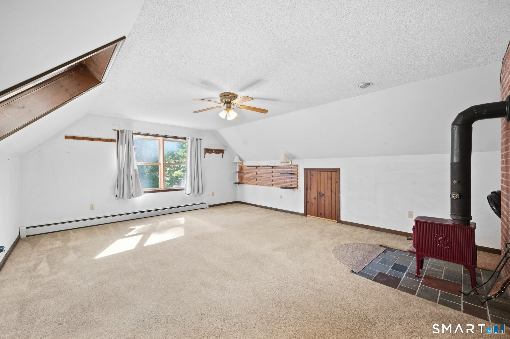 Empty vaulted room with beige carpet, a window, and a red wood stove on a tiled hearth.