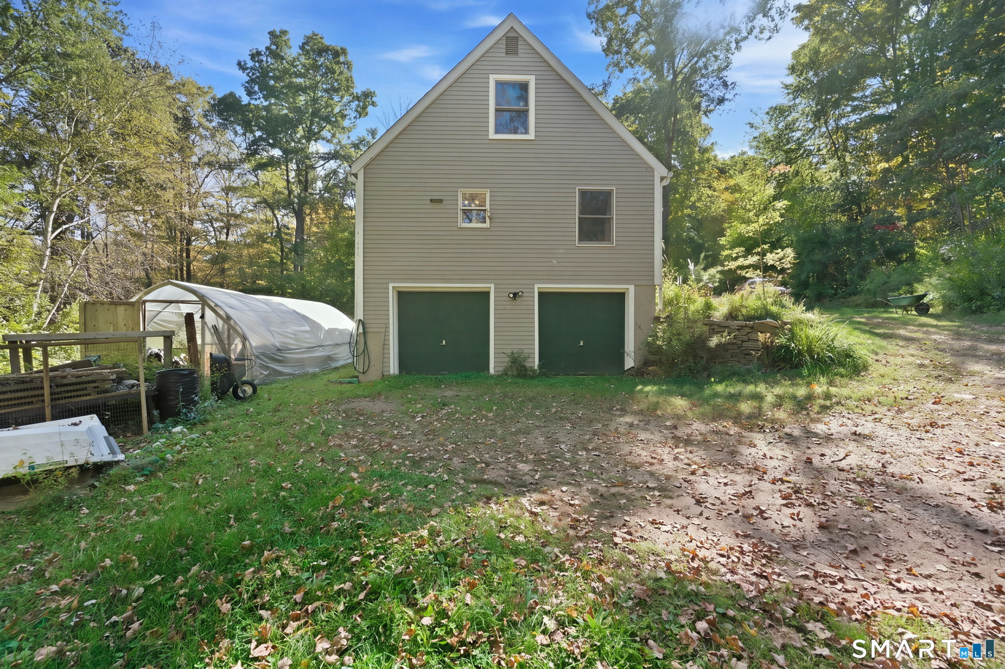 Gabled house with two green garage doors, wooded yard, and a covered garden structure.