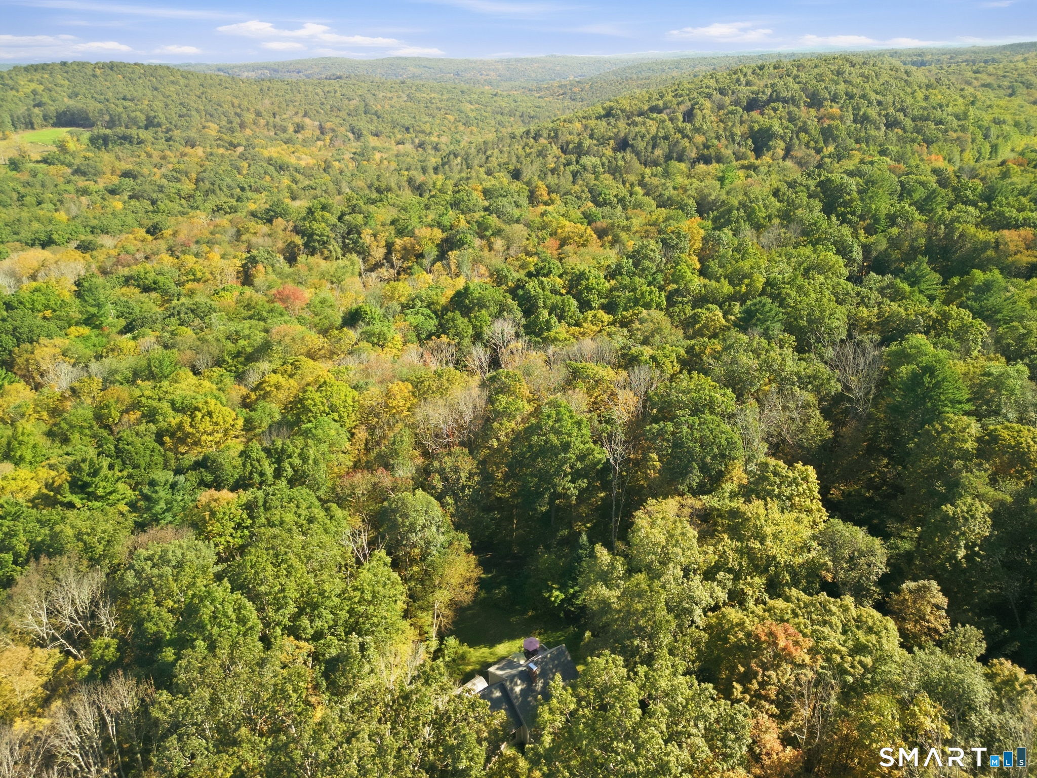 Aerial view of a house nestled in dense autumn woodland, with rolling forested hills beyond.