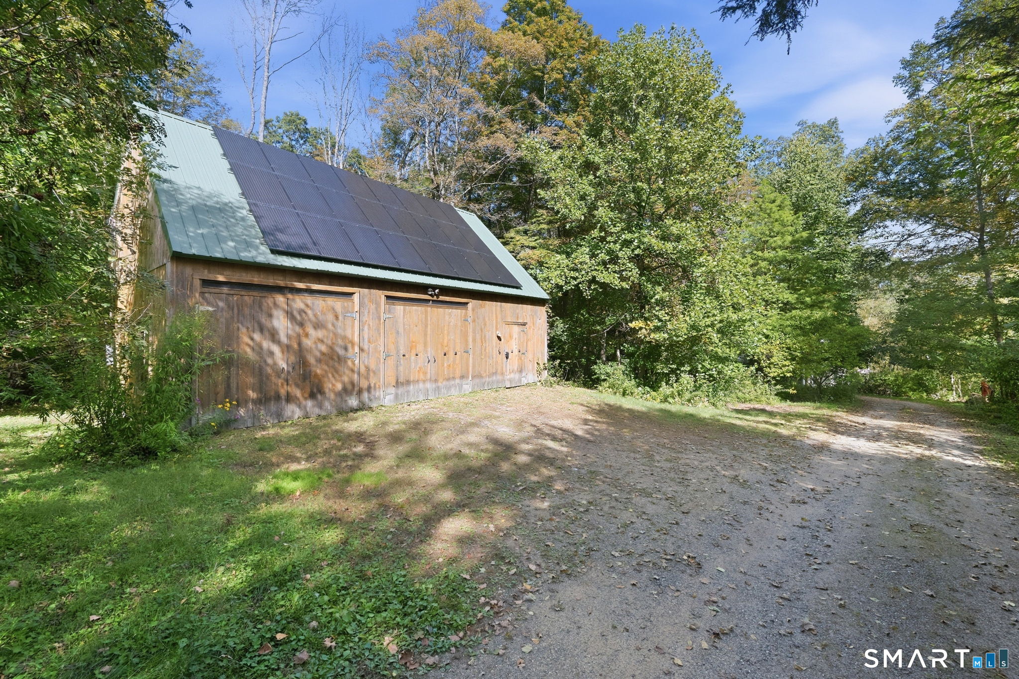 Wooden outbuilding with solar panels on its steep roof, beside a gravel drive in a wooded yard.