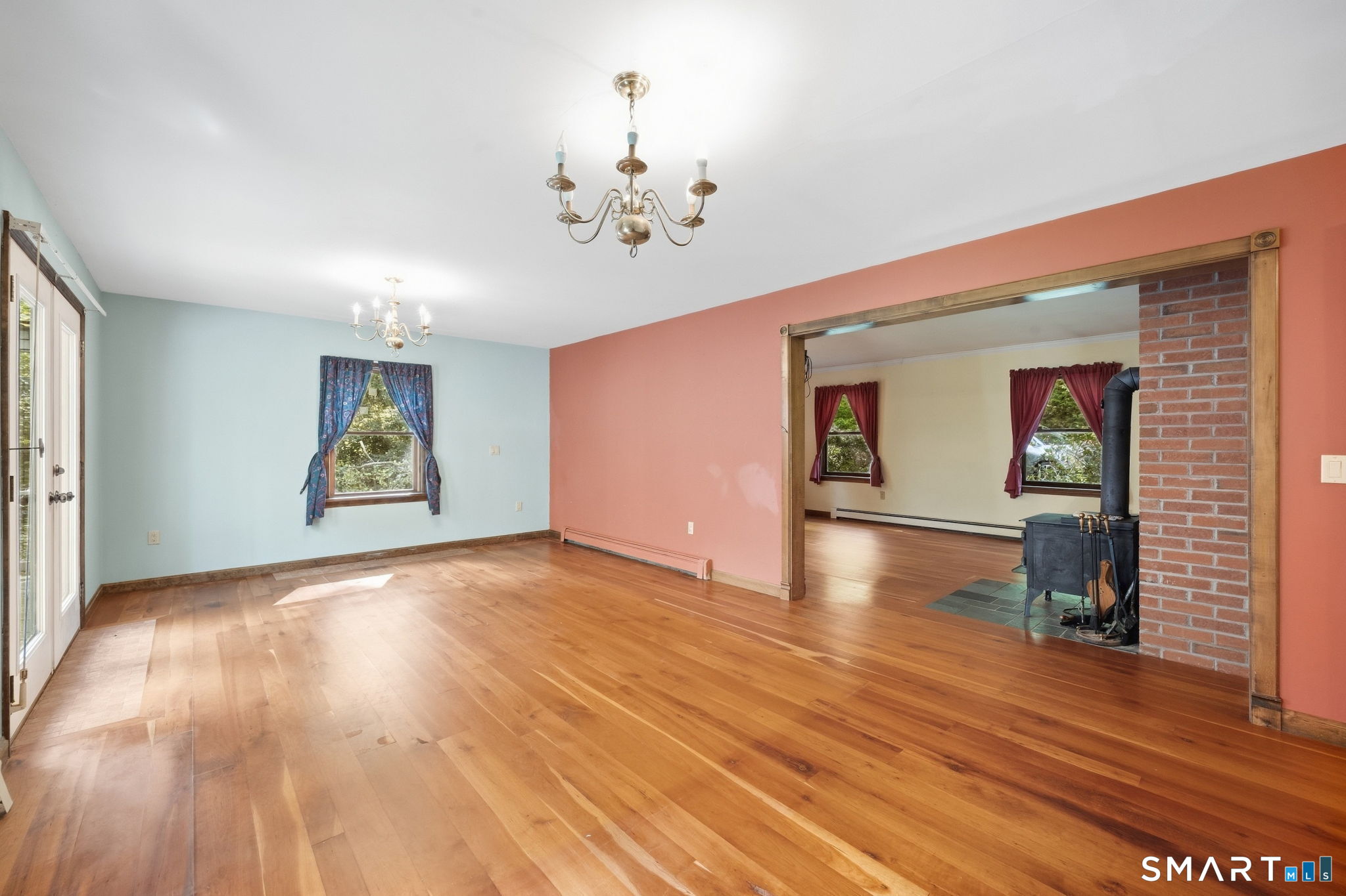 Living room with hardwood floors and chandeliers, opening to an adjoining room with a brick-backed wood stove.