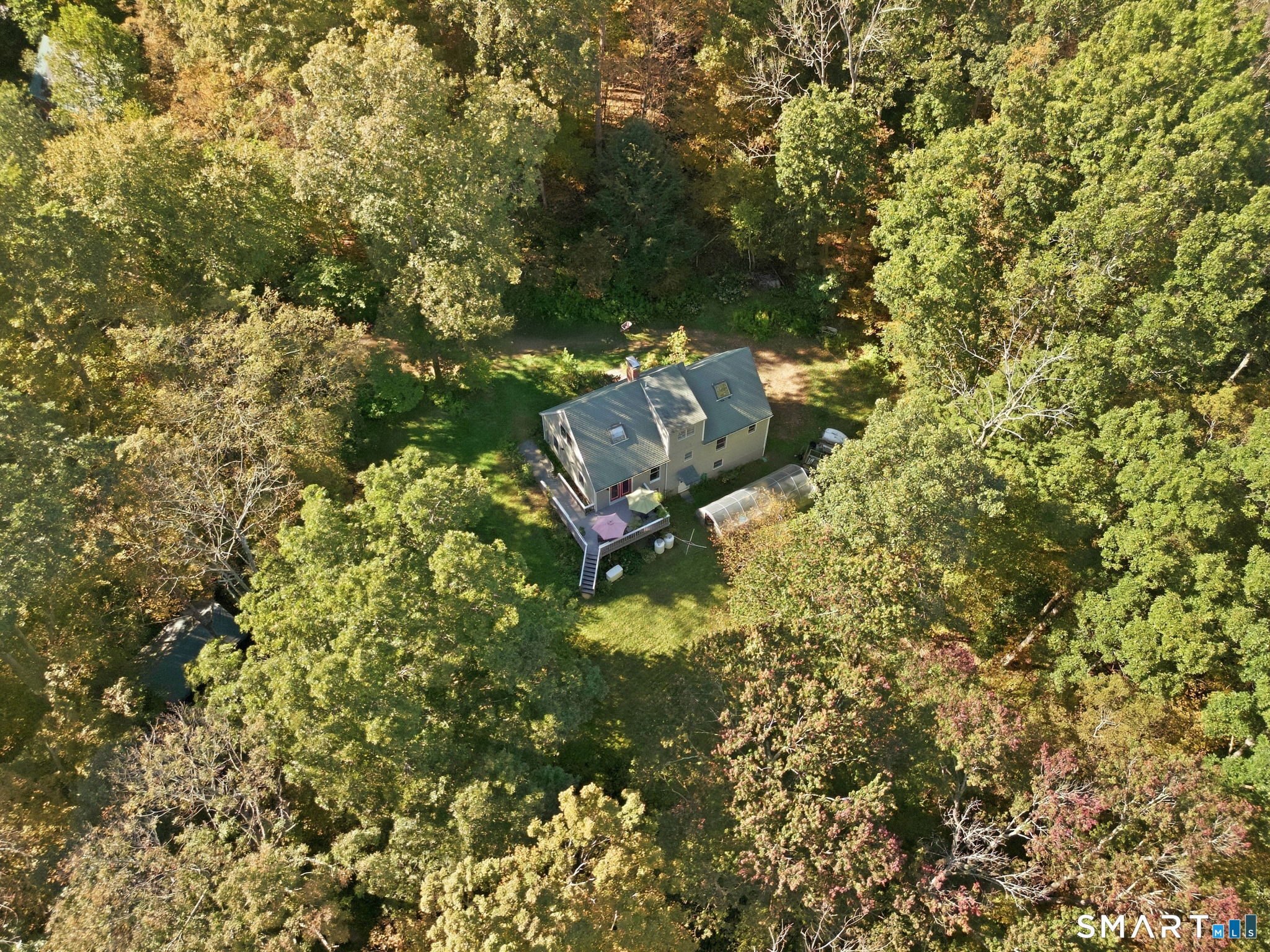 Aerial view of a home surrounded by trees, with a lawn, rear deck, and greenhouse.