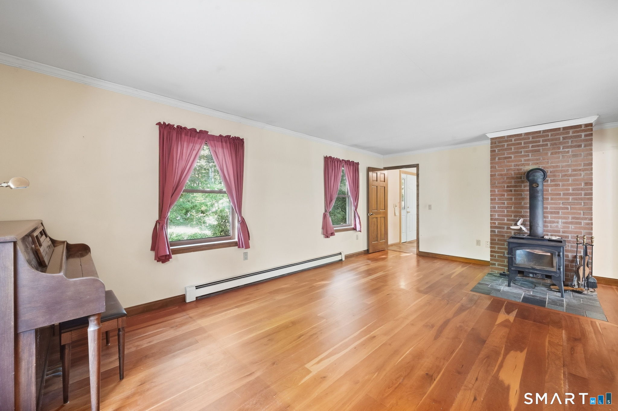 Living room with hardwood floors, a brick-surround wood stove, pink curtains, and a piano.