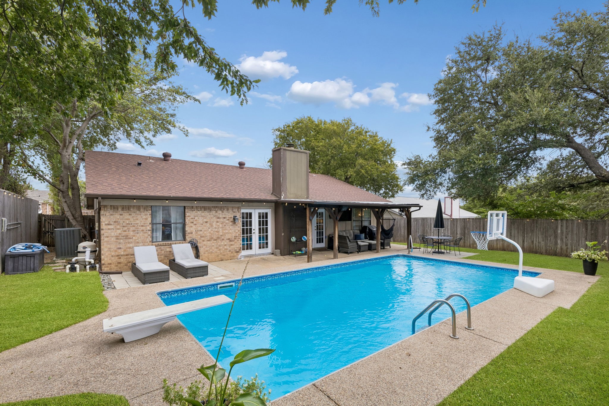 Brick home and covered patio behind a backyard swimming pool with a diving board and basketball hoop.