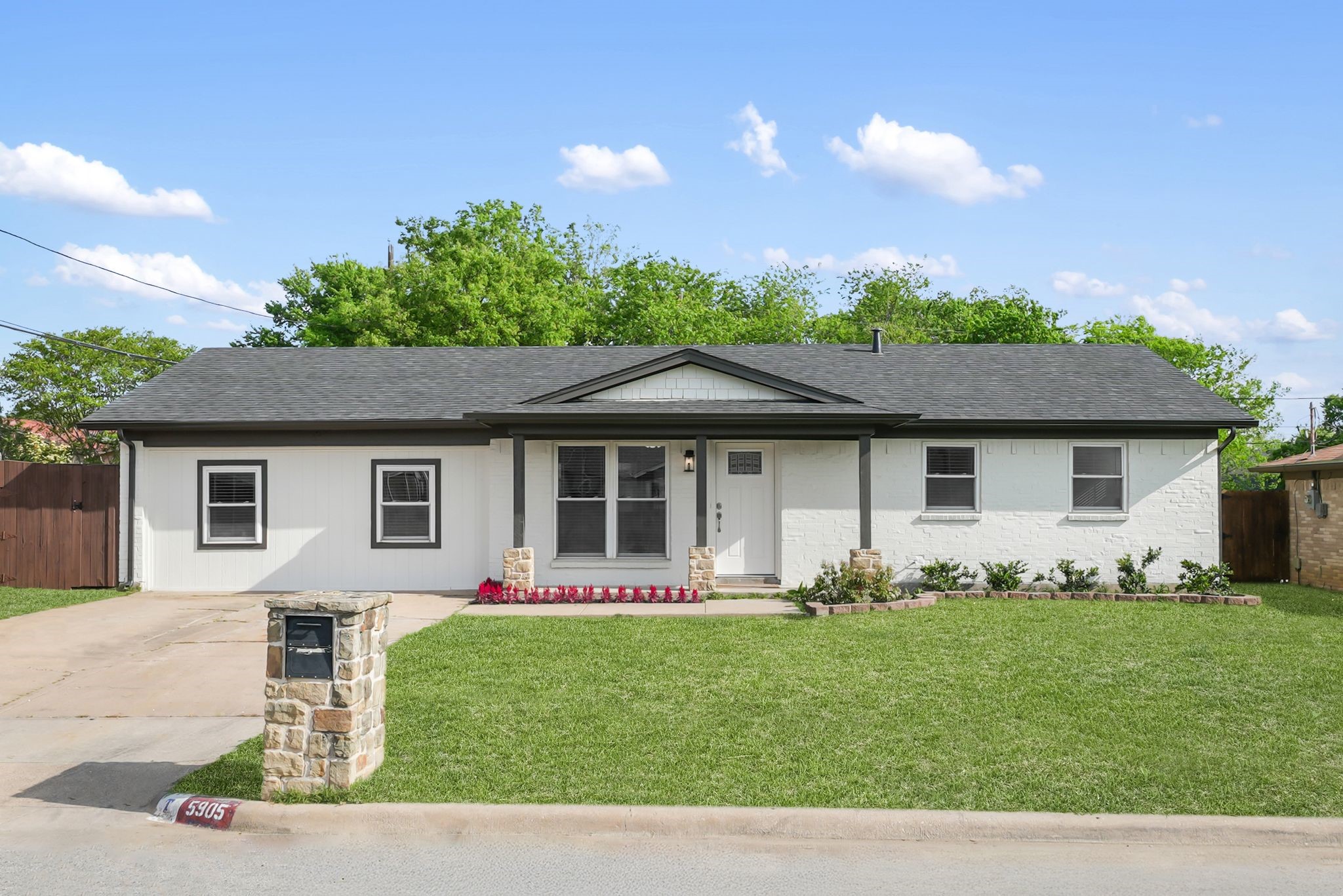 Single-story white brick house with a covered porch, broad lawn, driveway, and stone mailbox pillar.