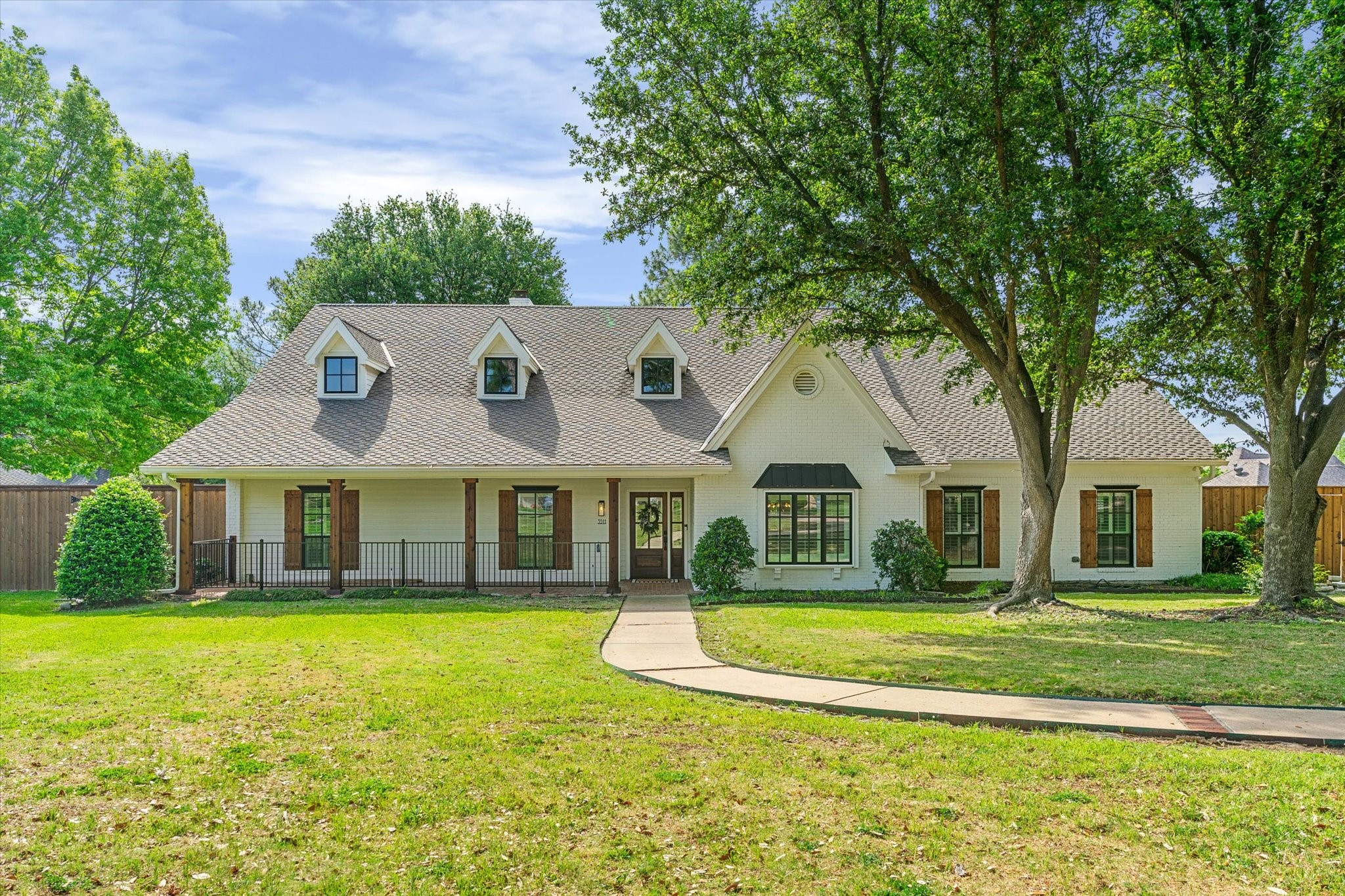 White brick house with three roof dormers, a covered porch, and large shade trees in the front yard.