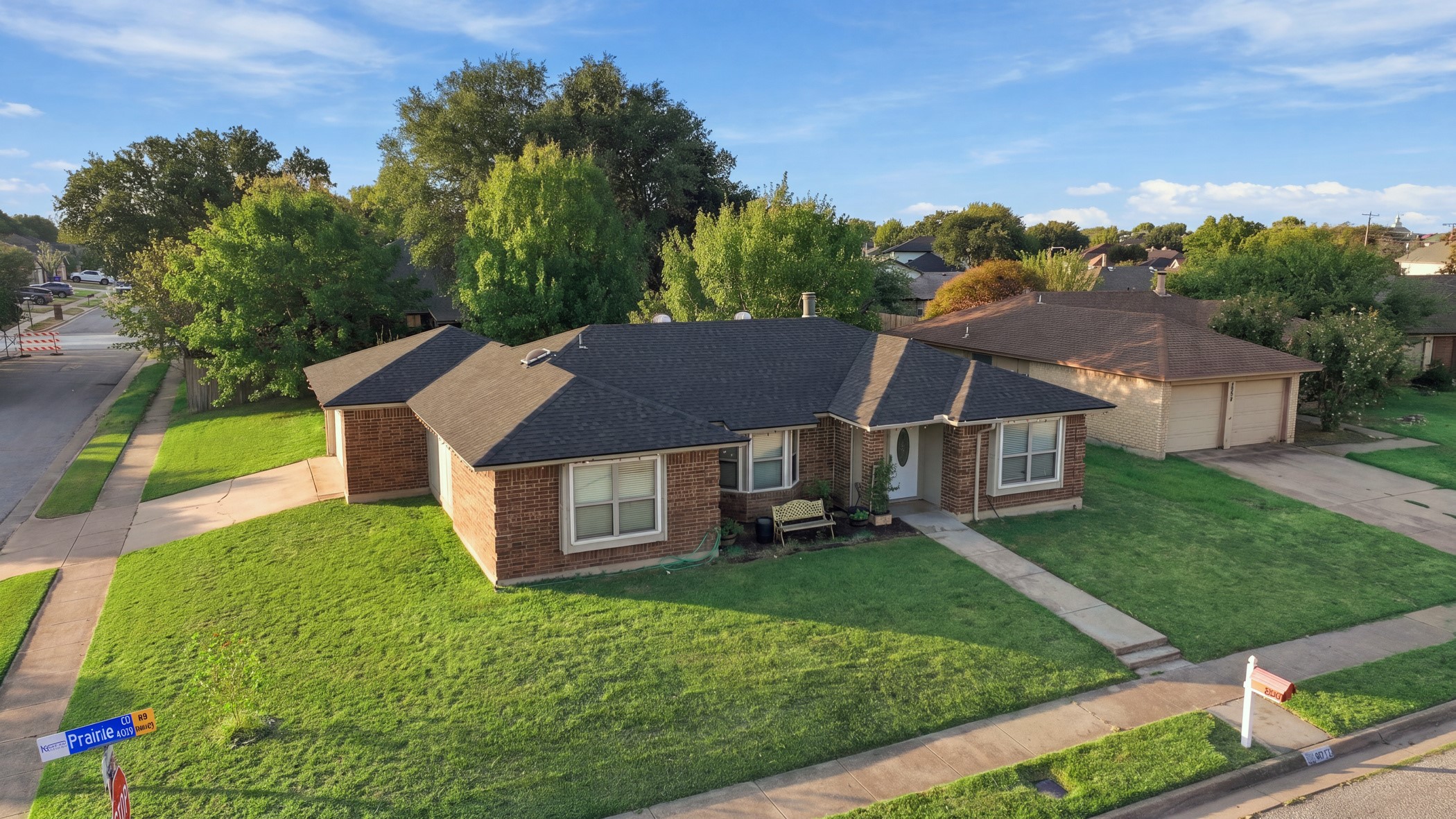 Aerial view of a brick ranch home with a broad green lawn and tree-lined neighboring homes.