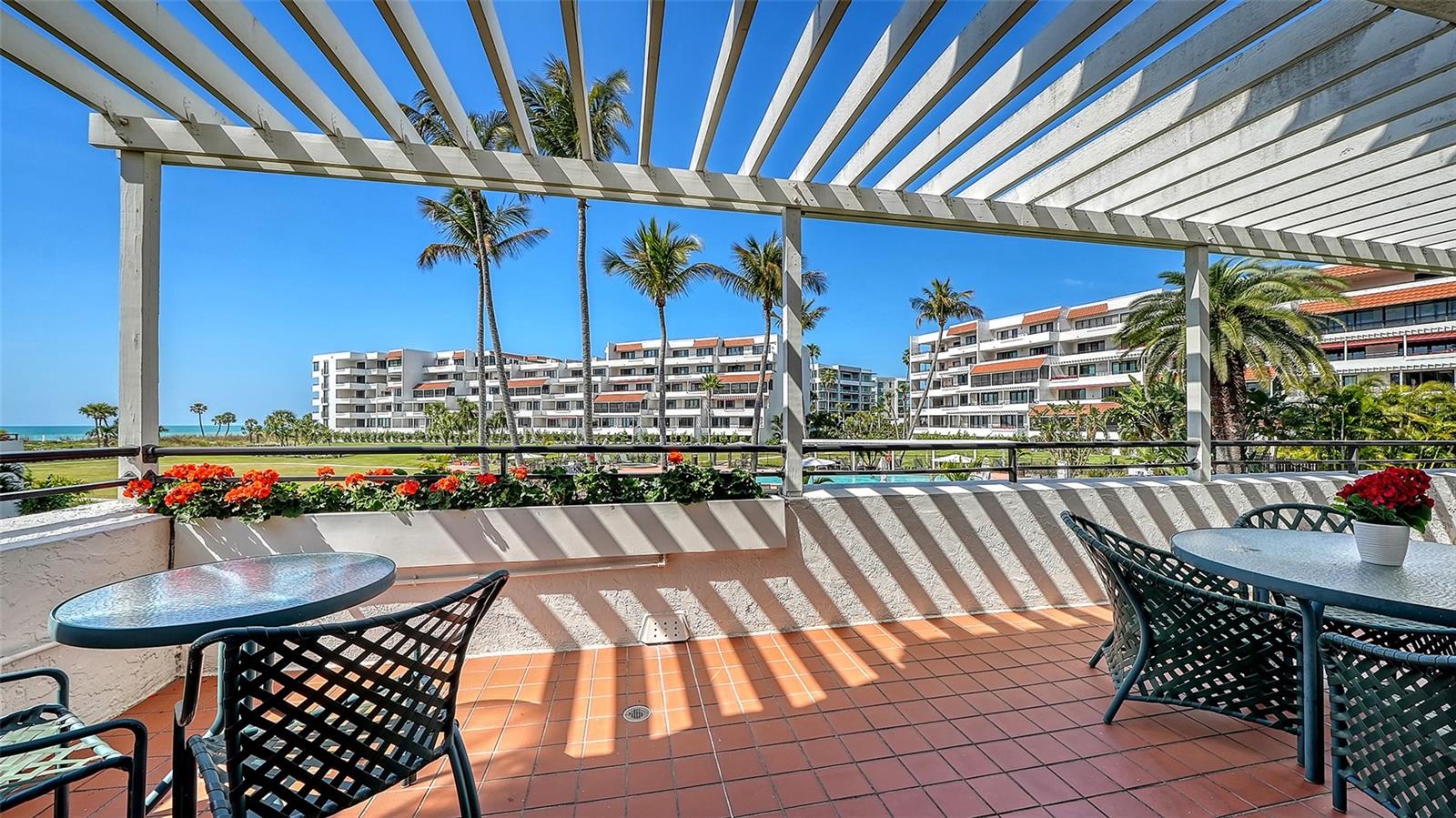 Tiled terrace with pergola, outdoor seating, flower planters, and views of palm-lined condominium buildings and ocean.
