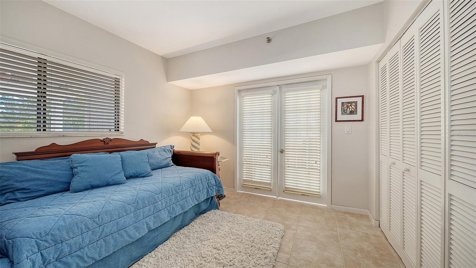 Bedroom with a blue bed, white louvered closet doors, and blinds covering the windows and glass doors.