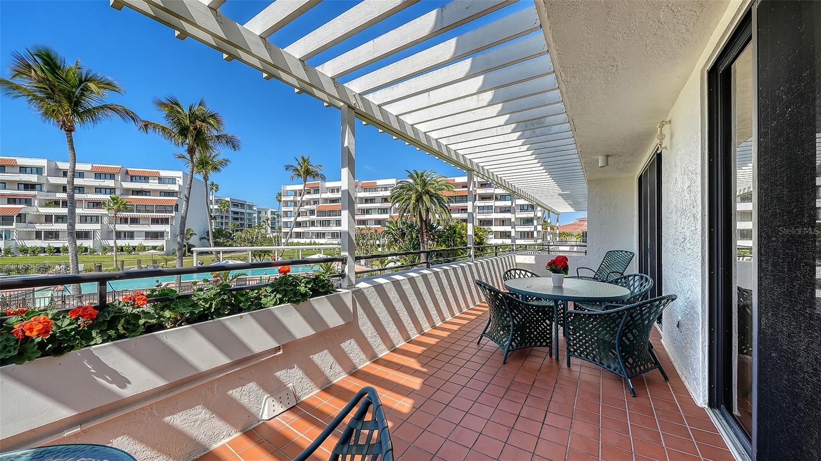 Furnished condo balcony with terracotta tiles, a white pergola, and views of the pool and palm-lined grounds.