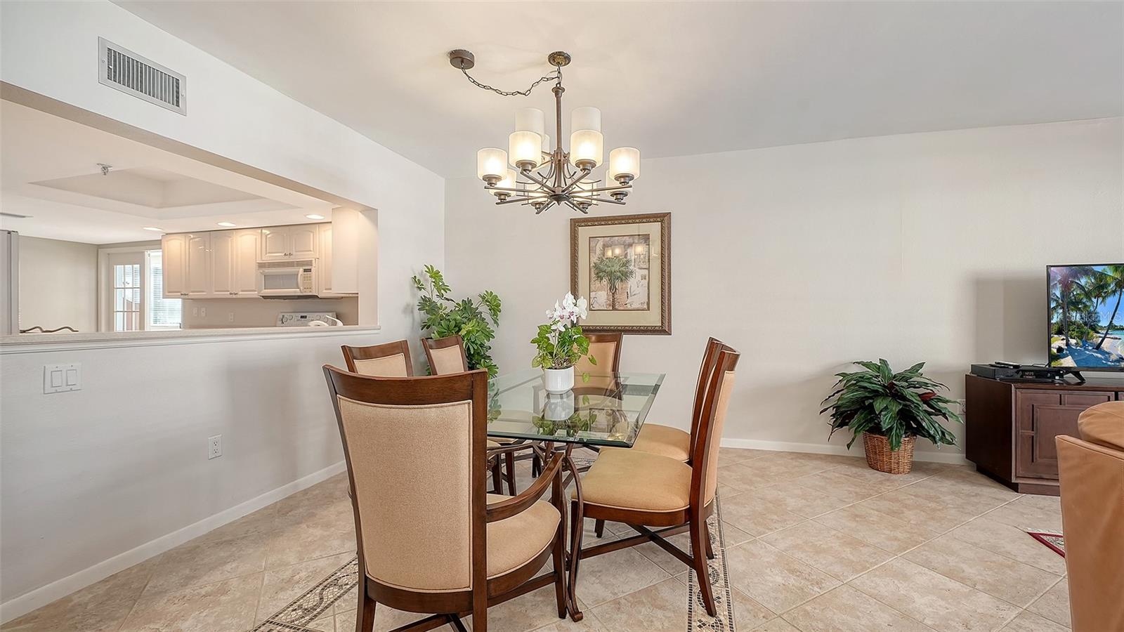 Dining area with a glass-top table, chandelier, and pass-through to the kitchen.