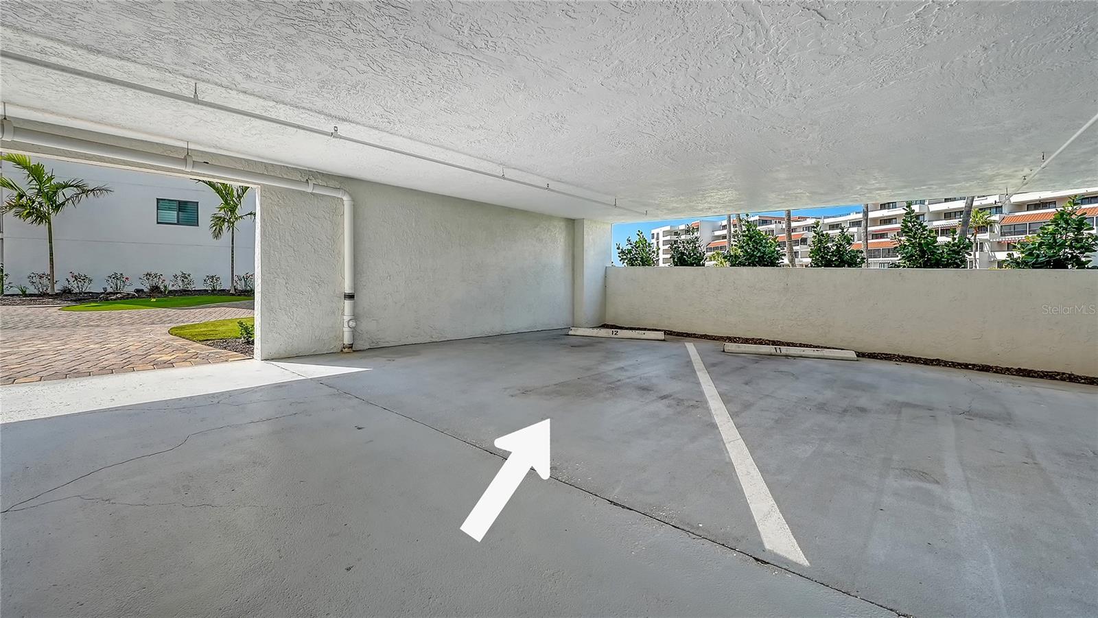 Covered parking space marked by a white arrow, with landscaping and condominium buildings beyond.