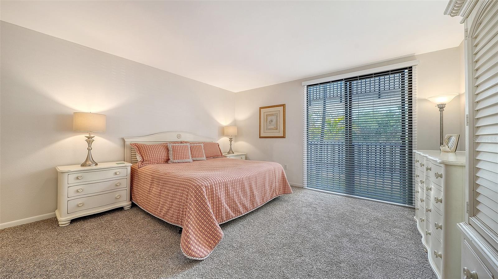 Furnished bedroom with a checked bedspread, gray carpet, white dressers, and a wide window with blinds.