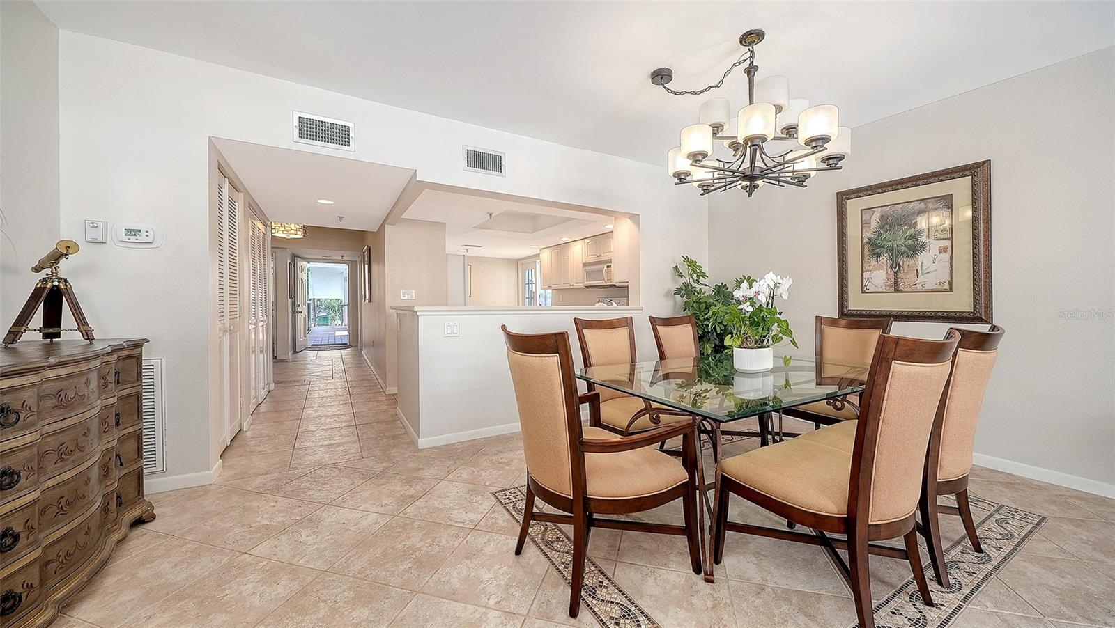 Dining area with a glass-top table, six upholstered chairs, chandelier, and tiled floor.
