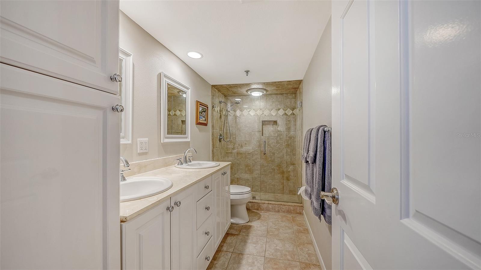 Bathroom with a double-sink vanity and large beige tiled walk-in shower.