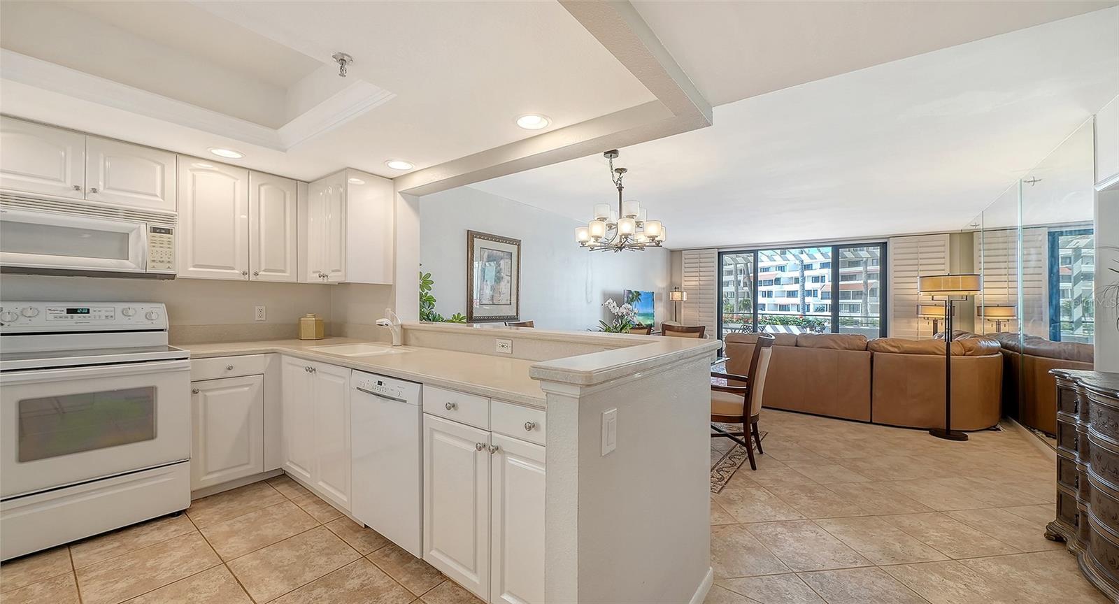 Condo kitchen with white cabinetry and peninsula opening to a living room with a brown leather sectional.