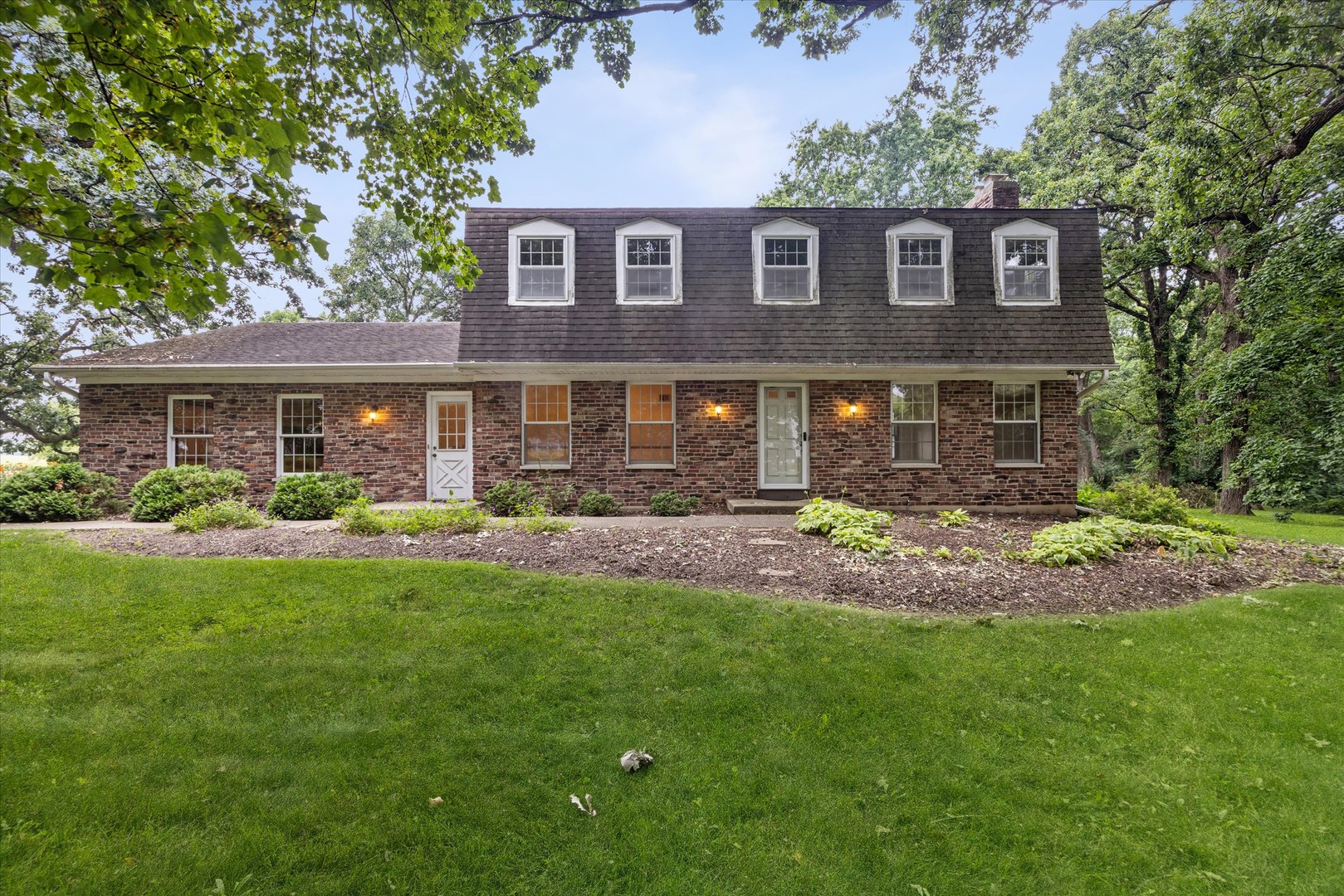 Brick house with five roof dormers, framed by mature trees and a broad green lawn.
