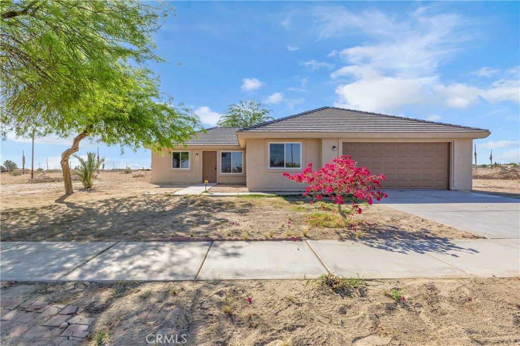 Single-story stucco home with an attached garage, shaded front yard, and bright pink flowering shrub.