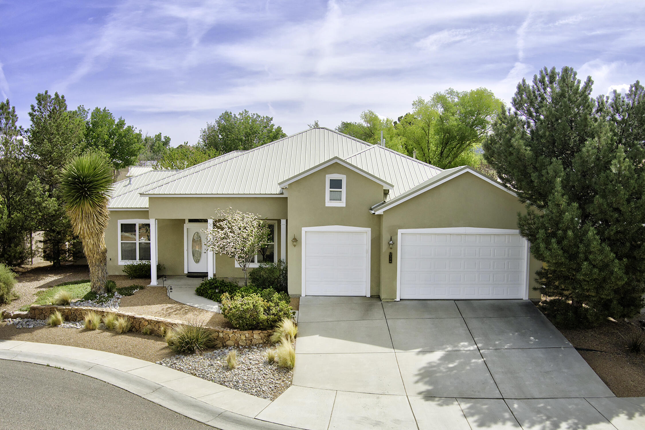 Tan stucco home with a white metal roof, two garages, and desert landscaping.