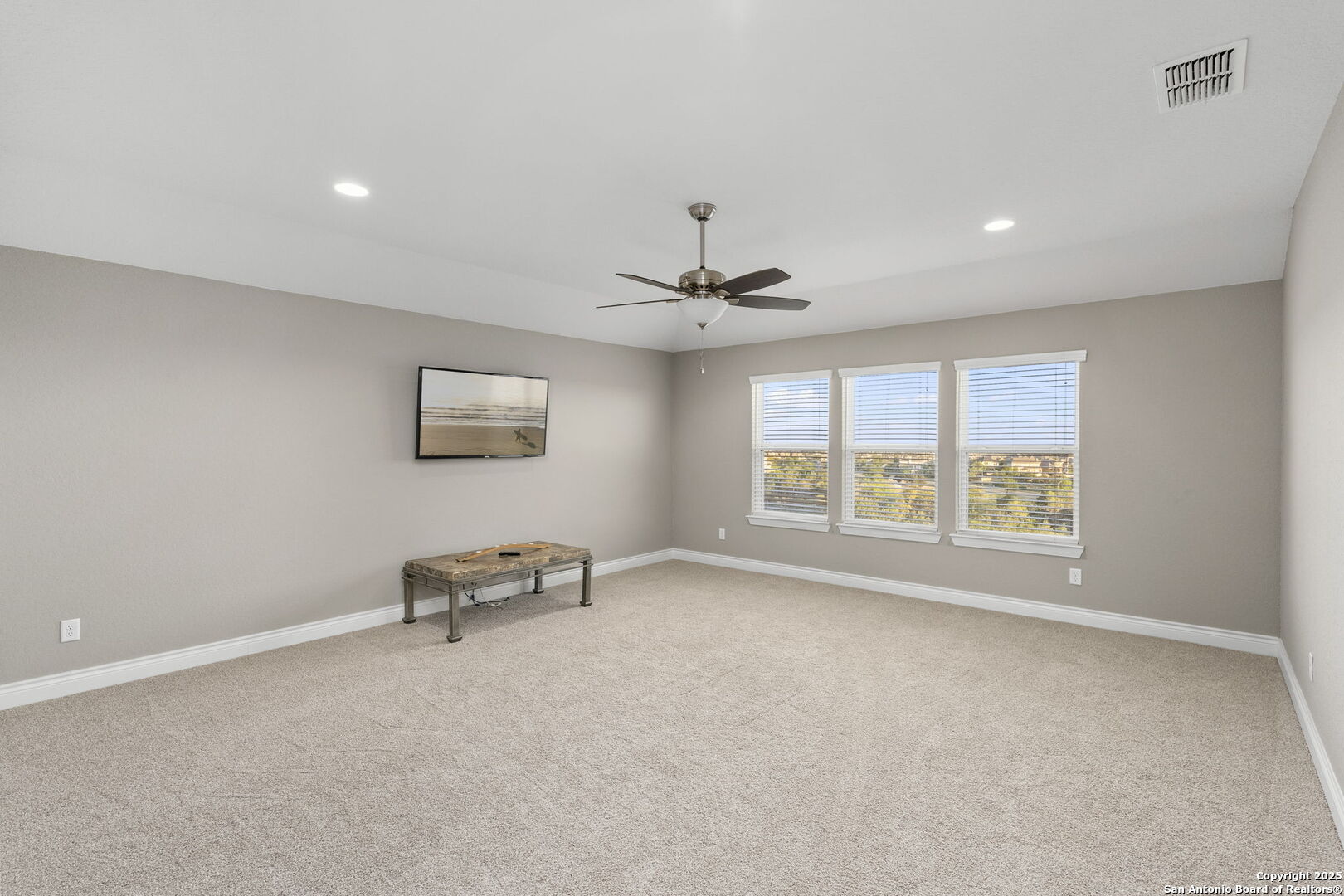 Spacious carpeted room with three windows, a ceiling fan, and a small table.