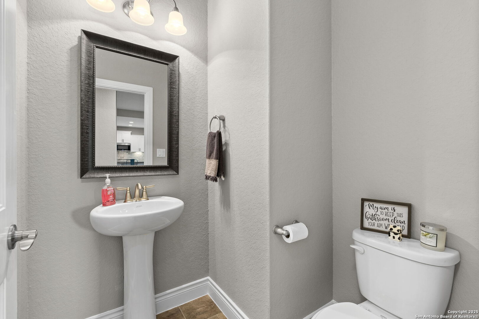 Bathroom with a white pedestal sink, dark-framed mirror, and toilet beneath a humorous cleaning sign.