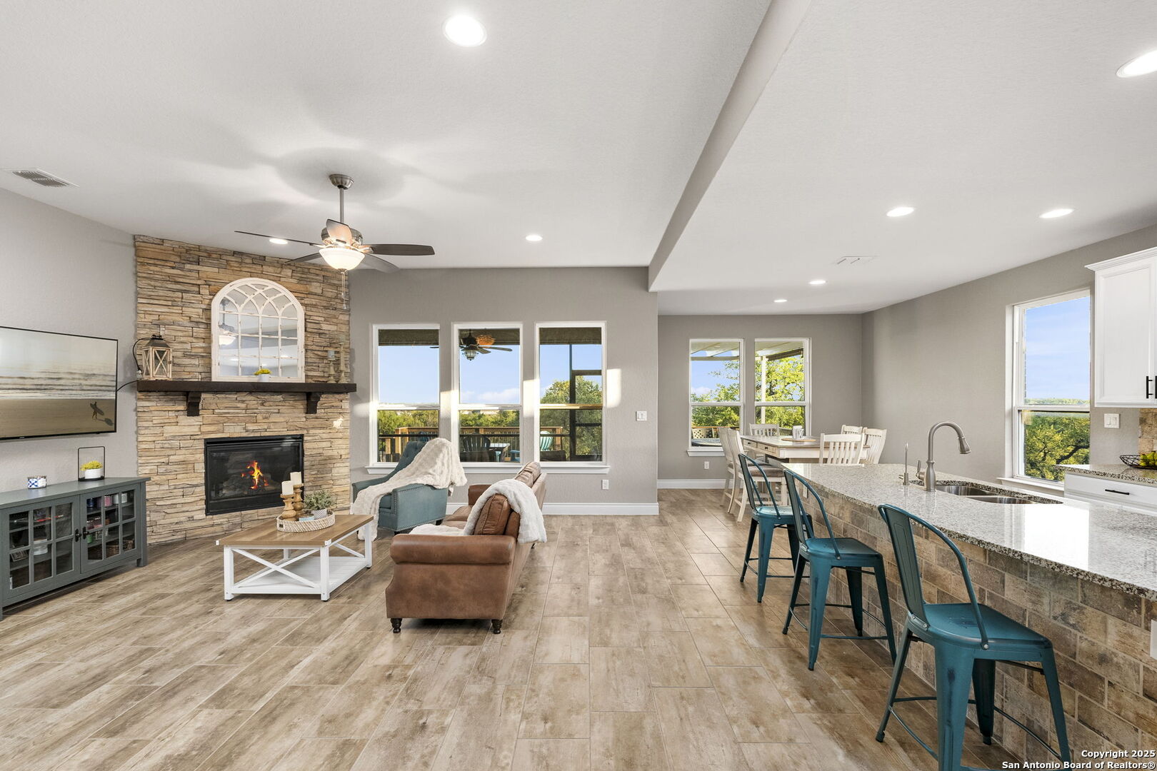 Open living room and kitchen with a stone fireplace, granite island, teal stools, and large windows.