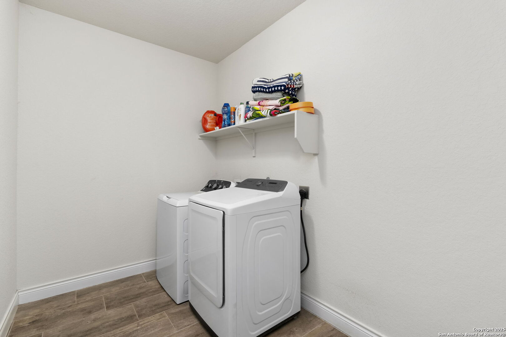 Laundry room with a white washer and dryer beneath a shelf of folded towels and supplies.