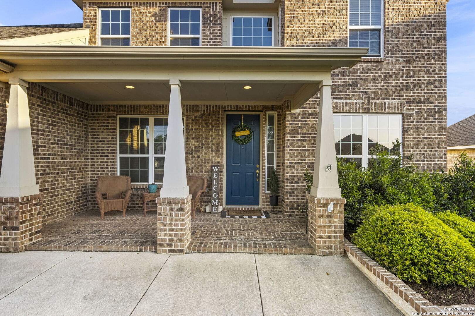 Two-story brick home with a covered porch, blue front door, wicker chairs, and landscaped shrubs.