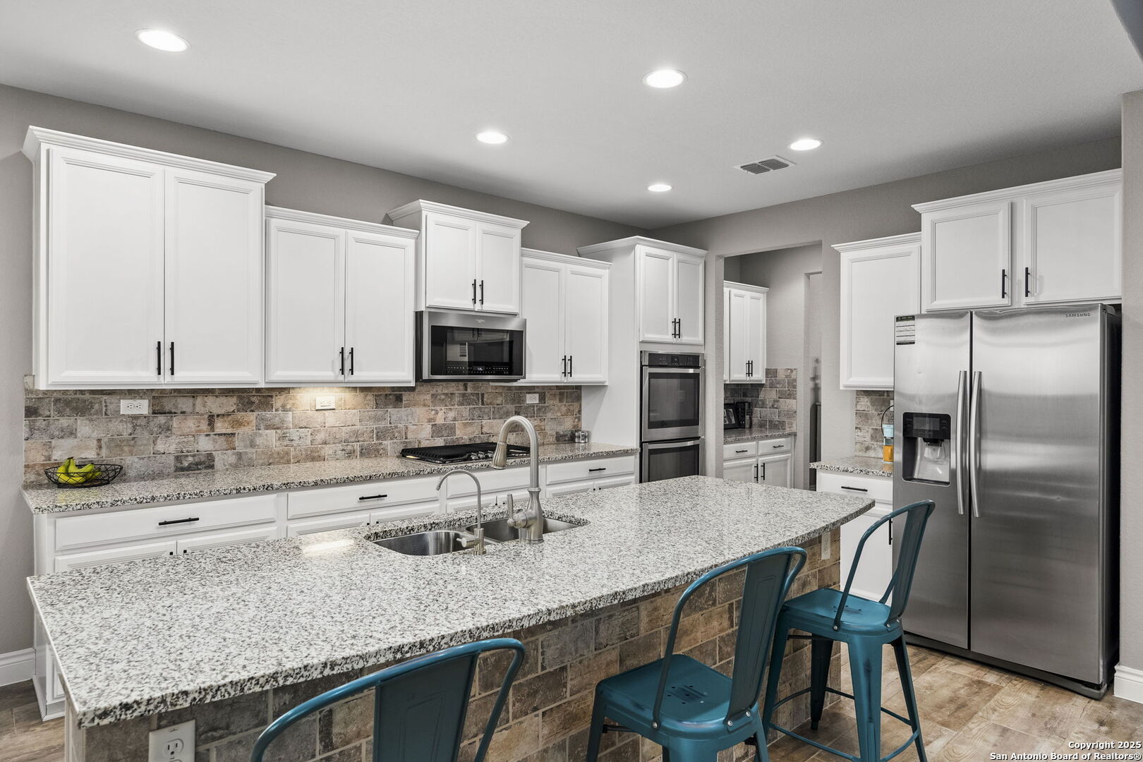 Kitchen with a large granite island, teal bar stools, white cabinetry, and stainless steel appliances.
