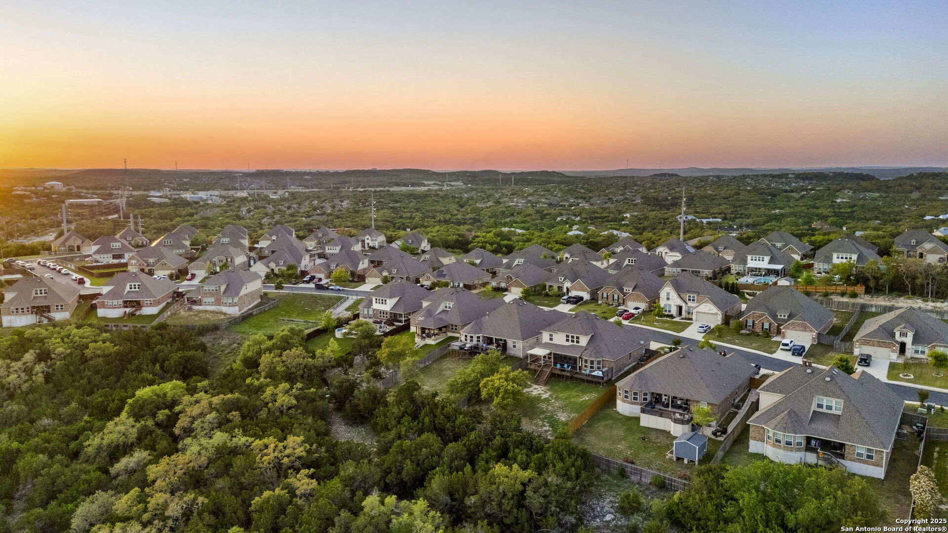 Aerial view of a residential neighborhood beside a wooded greenbelt under an orange dusk sky.